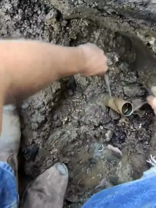 Through Jacob's glasses: hands working on an old supply pipe at the bottom of a dig
